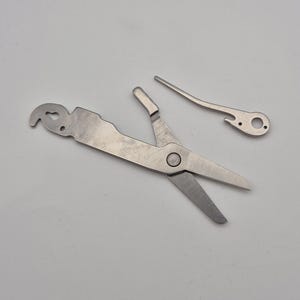 May include: A set of three silver-colored tools, including a pair of scissors, a curved lever, and a multi-tool with a hook and hole. The tools are arranged on a light gray surface, with the scissors open and the other pieces separate.