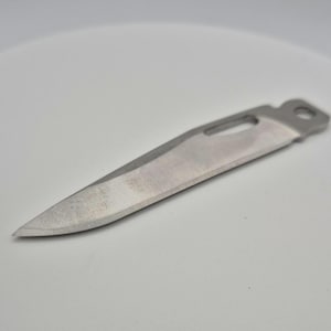 Op de afbeelding: Een zilverkleurig mesblad met een afgeronde punt en een klein gaatje bij het handvat.