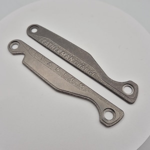 May include: Two grey titanium Leatherman Charge multi-tool accessories. One is engraved with "TITANIUM" and the other with "LEATHERMAN CHARGE". Each has a hole at one end and a curved design.