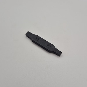 Peut inclure: Un outil rectangulaire noir avec le texte "R3" et "R2" imprimé dessus. L'outil a une forme légèrement en sablier avec des extrémités plus larges et une section centrale plus étroite. Le fond est gris clair uni.