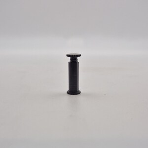 May include: A black, cylindrical object with a textured top and bottom. The object has a threaded design and appears to be made of metal. The background is a plain white surface.