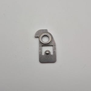May include: A small, silver-colored metal component with a unique shape. It features a circular hole near the top, a rectangular section with a rounded end, and a small, protruding circular element.