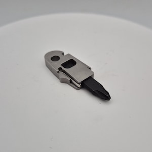 Puede incluir: Un componente multiherramienta compacto con un cuerpo de metal plateado que presenta un orificio circular en un extremo y un recorte rectangular. Una punta de destornillador Phillips negra sobresale del extremo opuesto, fijada en el marco.