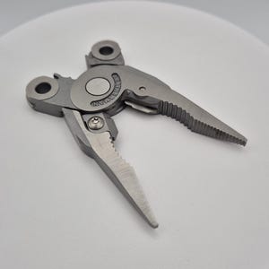 Puede incluir: Un cabezal de alicates multiherramienta Leatherman de tono plateado con un punto de pivote central que tiene el nombre de la marca grabado. La herramienta tiene dos mandíbulas dentadas y puntiagudas, un tornillo pequeño y dos puntos de anclaje.