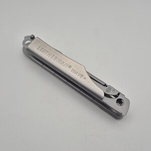 May include: A silver-colored stainless steel Leatherman Wave+ multi-tool in a closed, folded position. The metal handle features the engraved text LEATHERMAN WAVE+ and includes a small circular hole at one end and a locking mechanism detail.