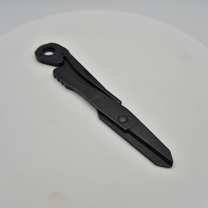 May include: A black, folding multi-tool with a key-shaped blade. The tool has a circular hole at the top and a riveted hinge. The tool is made of metal and has a matte finish.