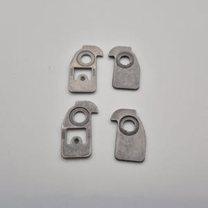 May include: Four small, silver-colored metal pieces. Each piece has a rounded top with a hole, a rectangular section, and a small hole. The pieces are arranged in a square formation against a white background.