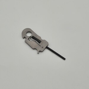 May include: A silver-toned metal tool featuring a circular opening at the top, a rectangular body with a spring-loaded wire latch mechanism, and a thin, straight black rod extending from the bottom edge of the metallic component.