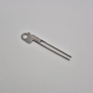 May include: A small, silver-coloured key-shaped tool. It has a circular opening at one end and two parallel prongs extending from the other. The tool is made of metal and appears to be a specialised instrument.