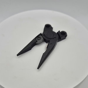 Pode incluir: Alicates pretos com uma mandíbula dentada e uma pega com mola. Os alicates são feitos de metal e têm um acabamento preto.