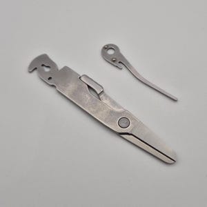 May include: Stainless steel scissors with a curved blade and a separate, smaller tool. The scissors have a folding design with a circular button. The smaller tool has a hook and a hole at the end.