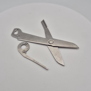 May include: A pair of small, silver-colored scissors with a separate, curved metal piece. The scissors have sharp blades and a loop at the end of one handle. The metal piece has a circular opening and a curved arm.