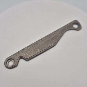 Pode incluir: Uma pequena ferramenta de titânio cinza com a palavra "TITANIUM" gravada ao longo do seu comprimento. A ferramenta tem uma forma curva com um furo em cada extremidade e uma superfície texturizada.