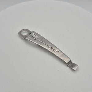 May include: A silver metal tool with a hole in the top and a curved end. The tool is labelled "LEATHERMAN®".