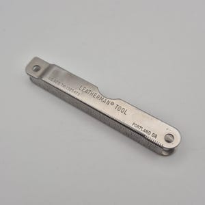 May include: A silver Leatherman Tool, featuring a ruler with inch markings. The tool has a hole at each end and the text "LEATHERMAN TOOL PORTLAND OR" is printed on the surface. The tool is registered under US REG TM 1325473.