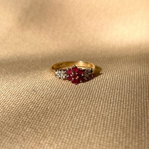 May include: A gold ring featuring a central cluster of red gemstones, framed by smaller white stones. The ring is displayed on a textured, neutral-toned surface, emphasizing the contrast and the ring's intricate design.