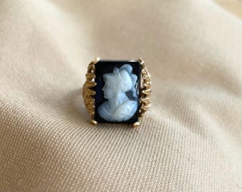 May include: A gold ring with a rectangular black cameo depicting a white female profile. The ring has a detailed, textured design around the cameo. The ring is set against a neutral background.