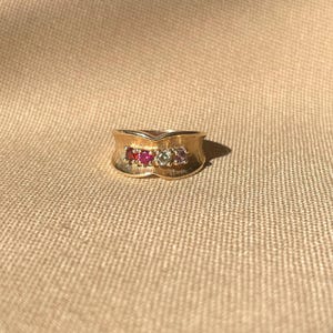 Könnte beinhalten: Ein goldener Ring mit strukturierter Oberfläche, mit einer Reihe von drei Edelsteinen: rot, rosa und klar. Der Ring hat ein einzigartiges, geschwungenes Design. Der Ring ist vor einem neutralen, strukturierten Hintergrund platziert.