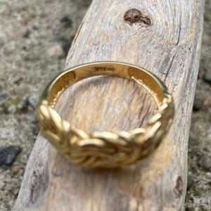 Puede incluir: Un anillo de color dorado con un dise&ntilde;o trenzado. El anillo est&aacute; sobre una superficie de madera.