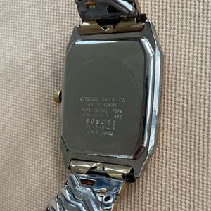 Peut inclure: Une montre Citizen argent&eacute;e et dor&eacute;e avec un cadran carr&eacute; et un bracelet en m&eacute;tal. Le dos de la montre porte l'inscription "CITIZEN WATCH CO. WATER RESIST BASE METAL FOFE 1000-885871 HSE 809213 28-0-845 MINT JAPAN".