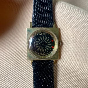 May include: Vintage gold-toned Borel Cocktail watch with a square face and a black dial featuring a floral design. The watch has a black leather band. The watch face has the word "Borel" and "Cocktail" printed on it.