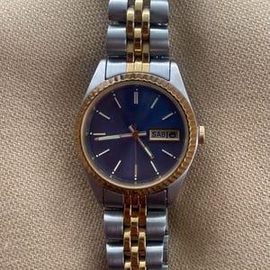 Puede incluir: Un reloj de pulsera de dos tonos, dorado y plateado, con una esfera azul y una pantalla del día de la semana. La esfera del reloj dice "SABI".