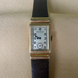 May include: A rectangular Bulova wristwatch with a silver face and black leather strap. The watch face displays the Bulova logo and black numerals. The watch has a gold-coloured case and a black leather strap.