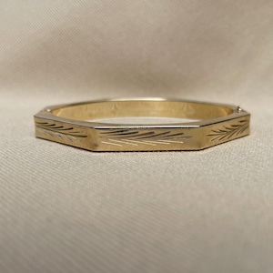 May include: A gold-toned metal bangle bracelet with a distinct hexagonal shape. The outer surface of the bangle features delicate, etched leaf-like patterns along its flat sides, providing a textured appearance to the polished metallic finish.