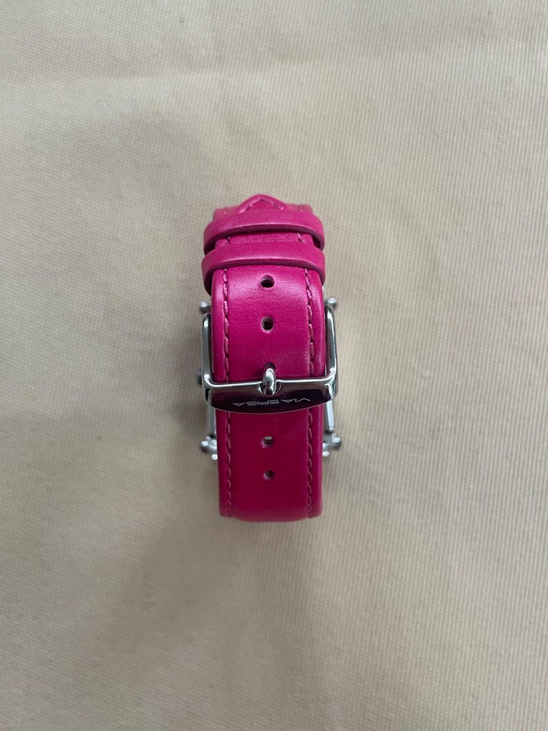Puede incluir: Una correa de reloj de cuero rosa con una hebilla plateada. La hebilla tiene la palabra "WISTER" grabada.