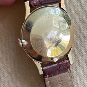 May include: Close-up of a vintage gold wristwatch with a brown leather band. The watch case is gold-toned with the marking "14K" visible. The band is a dark brown color.