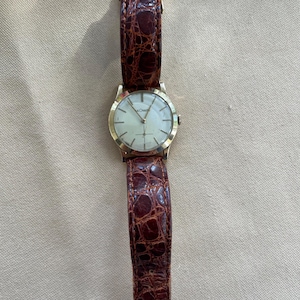 May include: A gold-toned wristwatch with a light-colored face and a brown leather band. The watch face has a simple design with thin hands and markers. The band has a textured, crocodile-like pattern.