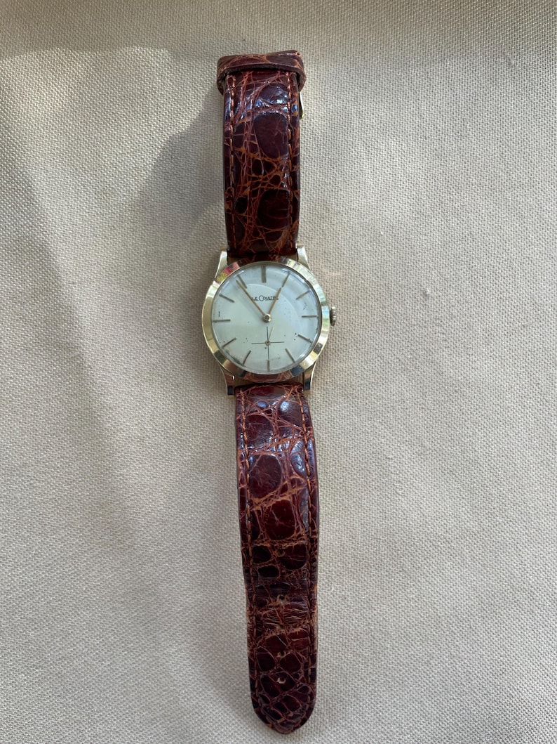 May include: A gold-toned wristwatch with a light-colored face and a brown leather band. The watch face has a simple design with thin hands and markers. The band has a textured, crocodile-like pattern.