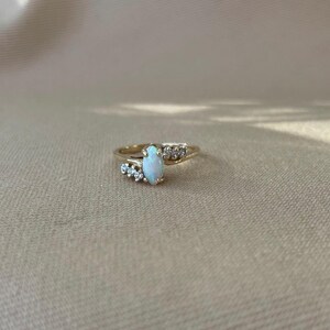 May include: A gold ring with a central oval opal stone and small clear accent stones. The ring has a delicate design, with the opal as the focal point. The ring is set against a neutral background.