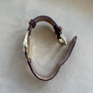 May include: A gold-toned wristwatch with a brown leather band. The watch face is circular, and the band is fastened with a gold-colored buckle. The watch is set against a neutral-toned background.