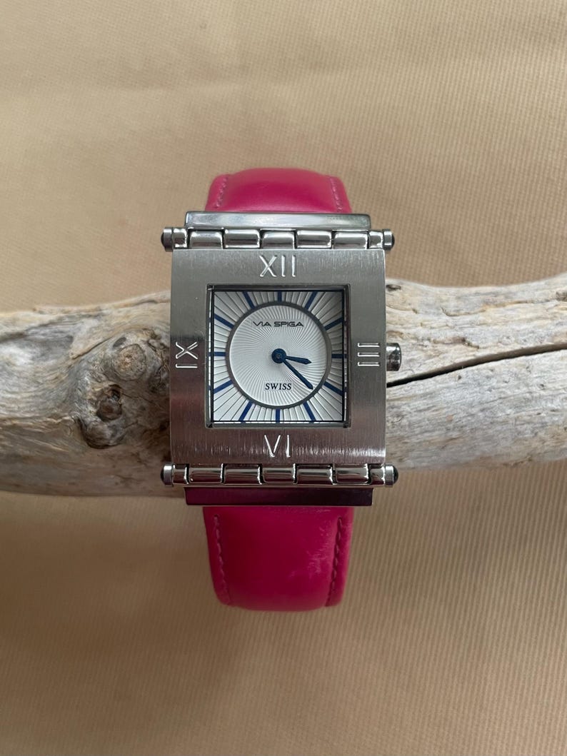 Puede incluir: Un reloj de pulsera cuadrado plateado con una esfera blanca y marcas de n&uacute;meros romanos negros. El reloj tiene una correa de cuero rosa y dice "Via Spiga" y "Swiss" en la esfera.