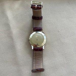 May include: A silver-toned wristwatch with a brown leather band. The watch has a round face and a buckle clasp. The band is a reddish-brown color. The word "INOX" is stamped on the clasp.