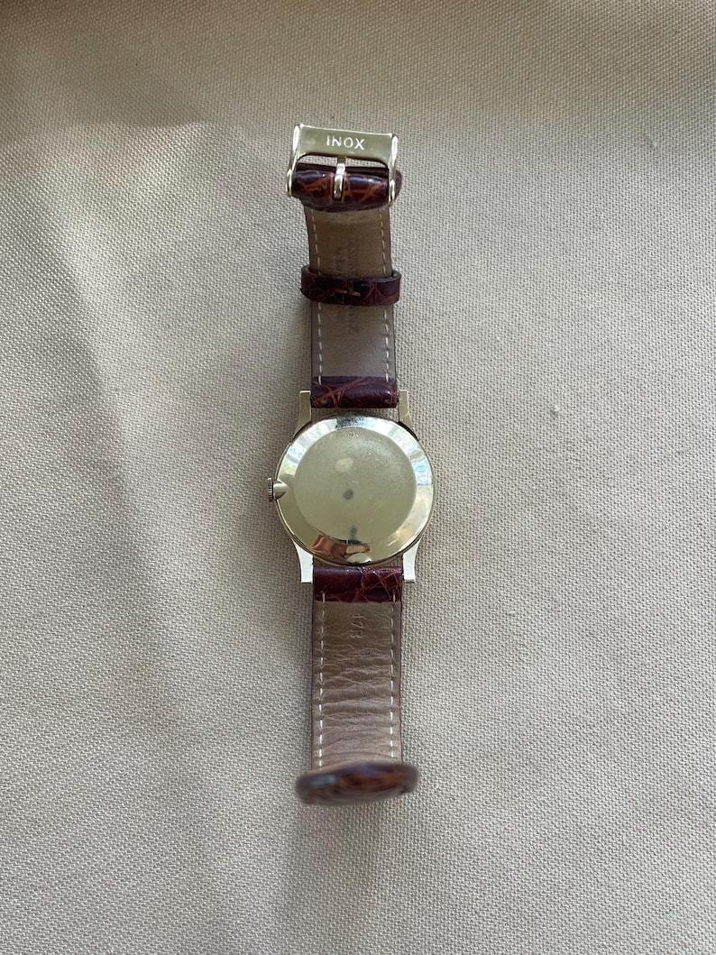 May include: A silver-toned wristwatch with a brown leather band. The watch has a round face and a buckle clasp. The band is a reddish-brown color. The word "INOX" is stamped on the clasp.