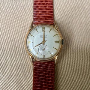May include: A vintage gold-toned wristwatch with a white face and gold hands and markers. The watch features the text "BENRUS 21 JEWELS". It has a brown leather strap with a textured pattern.
