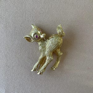 Könnte beinhalten: Goldfarbene Hirschbrosche mit strukturierter Oberfläche. Der Hirsch hat einen schlanken Körper, spitze Ohren und einen kleinen Schwanz. Die Augen sind mit kleinen, runden, roten Edelsteinen verziert. Die Brosche ist wahrscheinlich aus Metall und ein Vintage-Stück.