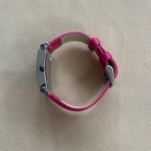 Puede incluir: Un reloj plateado con una esfera cuadrada y una correa de cuero rosa. El reloj tiene dos piedras preciosas azules en el lateral.