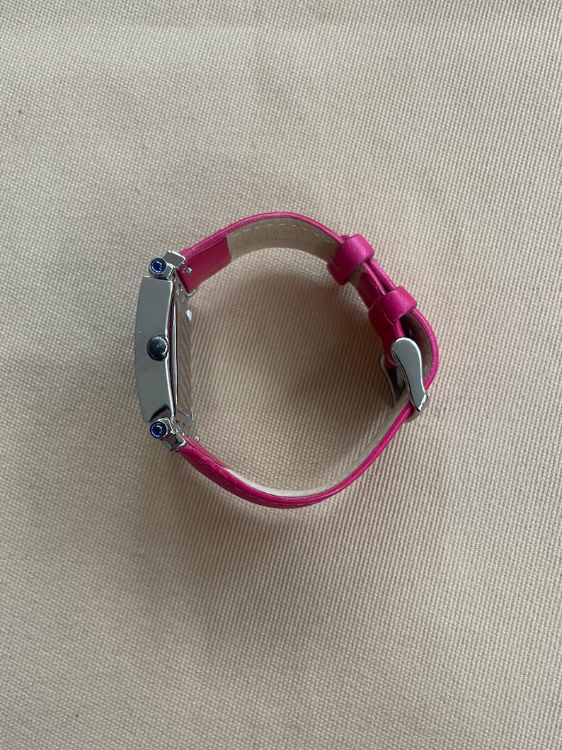 Puede incluir: Un reloj plateado con una esfera cuadrada y una correa de cuero rosa. El reloj tiene dos piedras preciosas azules en el lateral.