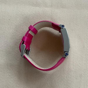 Puede incluir: Una correa de reloj de cuero rosa con una esfera de reloj plateada y detalles azules.
