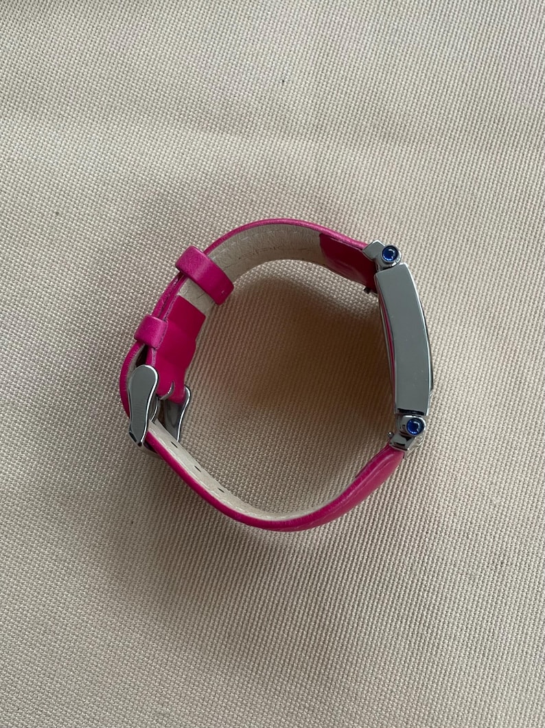 Puede incluir: Una correa de reloj de cuero rosa con una esfera de reloj plateada y detalles azules.