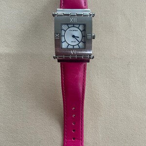 Puede incluir: Un reloj cuadrado plateado con una esfera blanca y n&uacute;meros romanos negros. El reloj tiene una correa de cuero rosa.