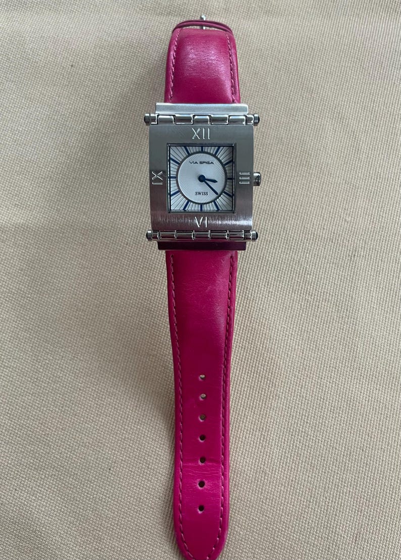 Puede incluir: Un reloj cuadrado plateado con una esfera blanca y n&uacute;meros romanos negros. El reloj tiene una correa de cuero rosa.