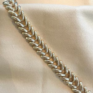 May include: A gold-toned bracelet with a herringbone design. The bracelet is composed of elongated, curved links that interlock to form a continuous chain. The clasp is visible at the end of the bracelet. The background is a light beige fabric.
