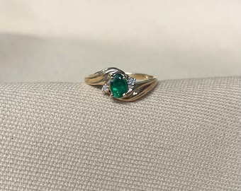 Peut inclure: Une bague en or ornée d'une émeraude ovale en son centre. La bague est ornée de petits diamants ronds de chaque côté de l'émeraude. Le corps de la bague présente un design fluide en forme de vague.