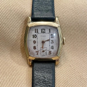 Könnte beinhalten: Eine vintage goldfarbene Picard-Armbanduhr mit rechteckigem Zifferblatt und schwarzem Lederarmband. Das Zifferblatt zeigt die Zahlen 1 bis 12 in einer klassischen Schriftart. Die Uhr hat eine kleine Sekundenanzeige und das Wort "SWISS" ist unten aufgedruckt.