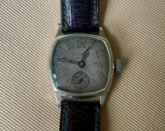 May include: A vintage wristwatch with a silver-toned case and a dark leather band. The watch face is a light gray with black numerals and hands. A smaller dial is at the bottom. The band is approximately 0.75 inches wide.