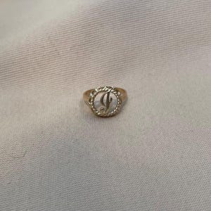 May include: A gold-toned ring featuring a circular centerpiece with a twisted rope-style border. The center of the ring displays a cursive letter J in a script font, set against a clear, open background within the circular frame.